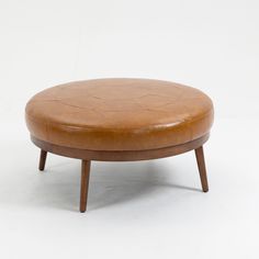 a round ottoman with wooden legs and a leather cushion on the top, against a white background