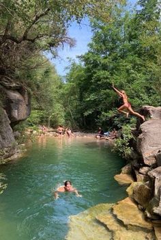 people are swimming in the water near some rocks and trees, while one person is jumping into