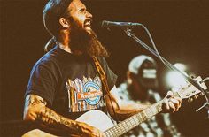 a man with a beard playing a guitar and singing into a microphone while another man looks on