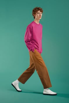 a young man in a pink shirt and brown pants walking across a green background wearing white tennis shoes