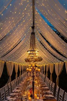 a long table with white chairs under a canopy covered in fairy lights and draping