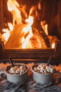 two mugs filled with hot chocolate and marshmallows sitting in front of a fireplace