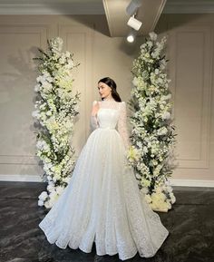 a woman standing in front of flowers wearing a wedding dress with long sleeves and lace