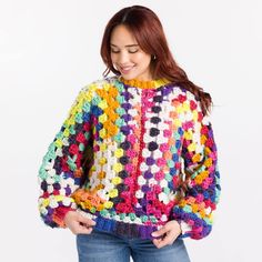 a woman wearing a multicolored crochet sweater and jeans, standing in front of a white background