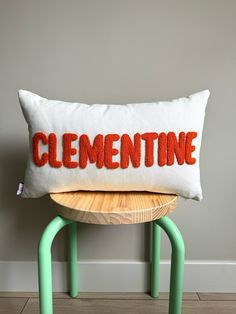 a white pillow with the word clemente on it sitting on a green stool in front of a gray wall