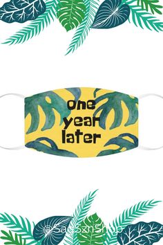 a face mask with the words one year later printed on it, surrounded by tropical leaves