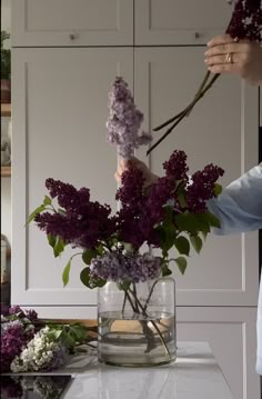 a person arranging flowers in a vase on a counter top with white cupboards behind them