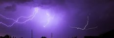 two lightning strikes in the night sky above power lines and trees, with purple hues