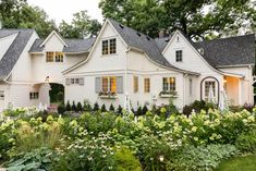 a white house surrounded by lots of flowers and greenery in front of the house