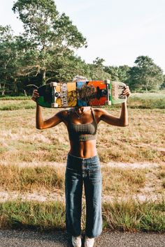 a woman holding a skateboard over her head in the middle of a field with grass and trees