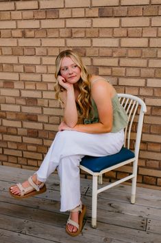 a woman sitting on top of a white chair next to a brick wall with her hand under her chin