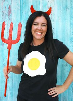 a woman wearing a black shirt with an egg on it and devil horns standing in front of a blue wall