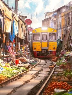 a train traveling through a market filled with lots of vegetables and fruit next to tall buildings