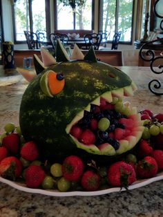 a watermelon shaped like a dinosaur with its mouth open and fruits in the foreground