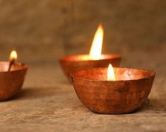 three copper bowls with lit candles in them on a stone surface, one is empty