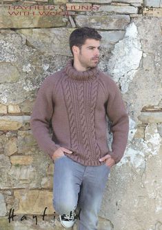 a man standing in front of a stone wall wearing a brown sweater and blue jeans