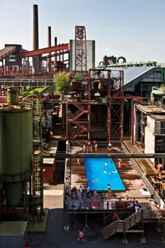 an overhead view of people playing in the water near industrial buildings and factory towers, with one swimming pool surrounded by metal pipes