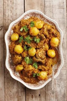 a bowl filled with meat and potatoes on top of a wooden table next to a spoon