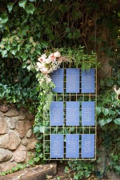 a blue sign with writing on it sitting next to some flowers and greenery in front of a stone wall