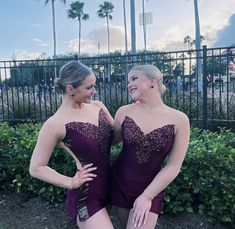 two women in short purple dresses standing next to each other near bushes and palm trees