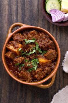 a brown bowl filled with meat and vegetables on top of a wooden table next to some napkins