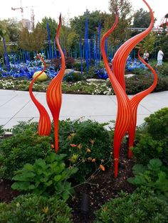 an orange sculpture is in the middle of some plants and flowers with blue glass sculptures behind it