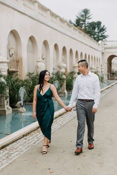 a man and woman holding hands walking in front of a fountain