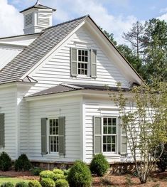 a white house with two windows and a steeple on the roof is surrounded by shrubs