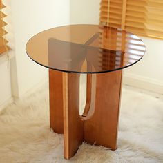 a glass table with wooden legs on a white rug in front of blinds and windows