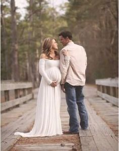 a pregnant couple standing on a bridge in the woods with their arms around each other