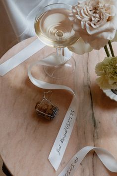 a table topped with a wine glass filled with liquid and flowers next to a white ribbon