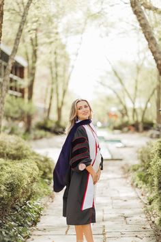 a woman wearing a graduation gown standing on a sidewalk in front of trees and bushes