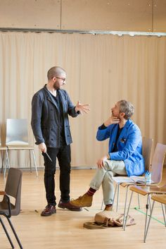 two people standing and sitting in chairs talking to each other