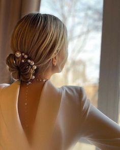 a woman with her back to the camera wearing pearls in her hair, looking out a window