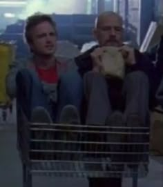 two men sitting in a shopping cart and one is holding a hot dog while the other looks on