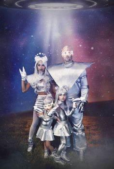 three people dressed in silver standing next to each other with an alien like object above them