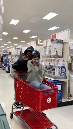 two people sitting in a red shopping cart taking pictures with their cell phones at the store