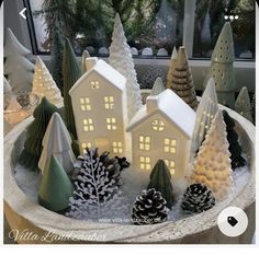 lighted christmas trees and small houses in a bowl on a table with a window behind them