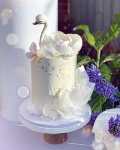 a white cake with flowers and a swan on top is sitting on a table next to purple flowers