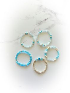 Blue Evil Eye protective Ring Set Chose your size US size 4-13  5 stretchy beaded rings.  Rings are made of a stretchy material with iridescent blue, light blue , and gold  premium glass seed beads plus one tiny evil eye protective bead , one light blue glass bead , and a cube white glass bead.  Handmade,  Perfect summer accessories ! Custom sizes available . Adjustable Light Blue Jewelry With Spacer Beads, Trendy Inelastic Round Beaded Jewelry, Blue Stackable Jewelry With Round Beads, Adjustable Stackable Rings With Tiny Beads As Gifts, Adjustable Beaded Stackable Rings, Blue Beaded Rings As Gift, Blue Beaded Rings As A Gift, Blue Beaded Rings As Gifts, Blue Beaded Rings For Gifts