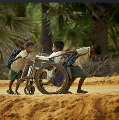 two boys pushing a wheel chair on the side of a dirt road with palm trees in the background