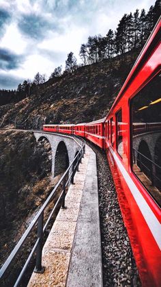 a red train traveling over a bridge next to a lush green hillside on a cloudy day