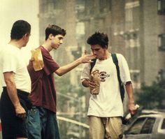 three young men standing on the side of a road looking at their cell phones and talking to each other