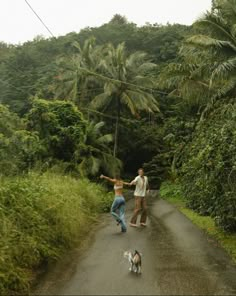 two people walking down a dirt road with a dog on one side and palm trees in the background