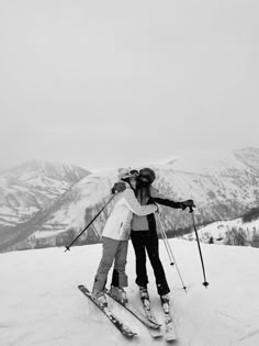 two women standing on top of a snow covered slope wearing skis and holding their arms around each other