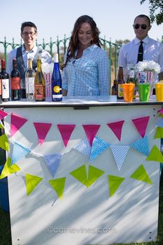 two men and a woman are standing behind a bar with drinks on it, in the grass