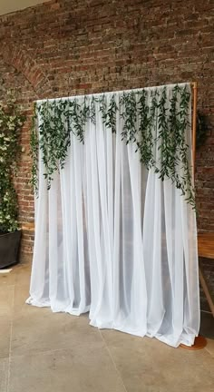 a white curtain with green plants on it in front of a brick wall and wooden bench