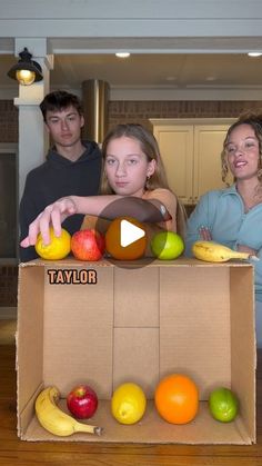 three people standing in front of a cardboard box filled with fruit