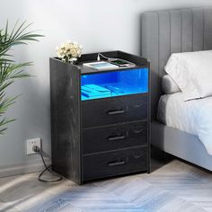 a night stand with three drawers in front of a bed and plant on the side