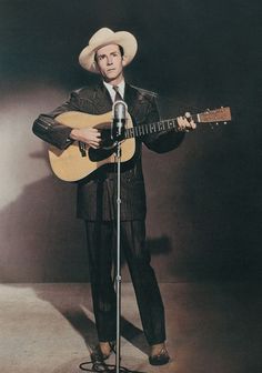 a man wearing a cowboy hat and holding a guitar while standing in front of a microphone